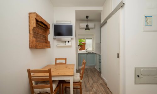 Sala de refeições do estudio T0 que dispõe de uma mesa de madeira e duas cadeiras, e que liga a cozinha. Por cima da mesa de refeição, tem uma televisão fixa na parede, uma peça decorativa em madeira, e uma porta de correr à direita.